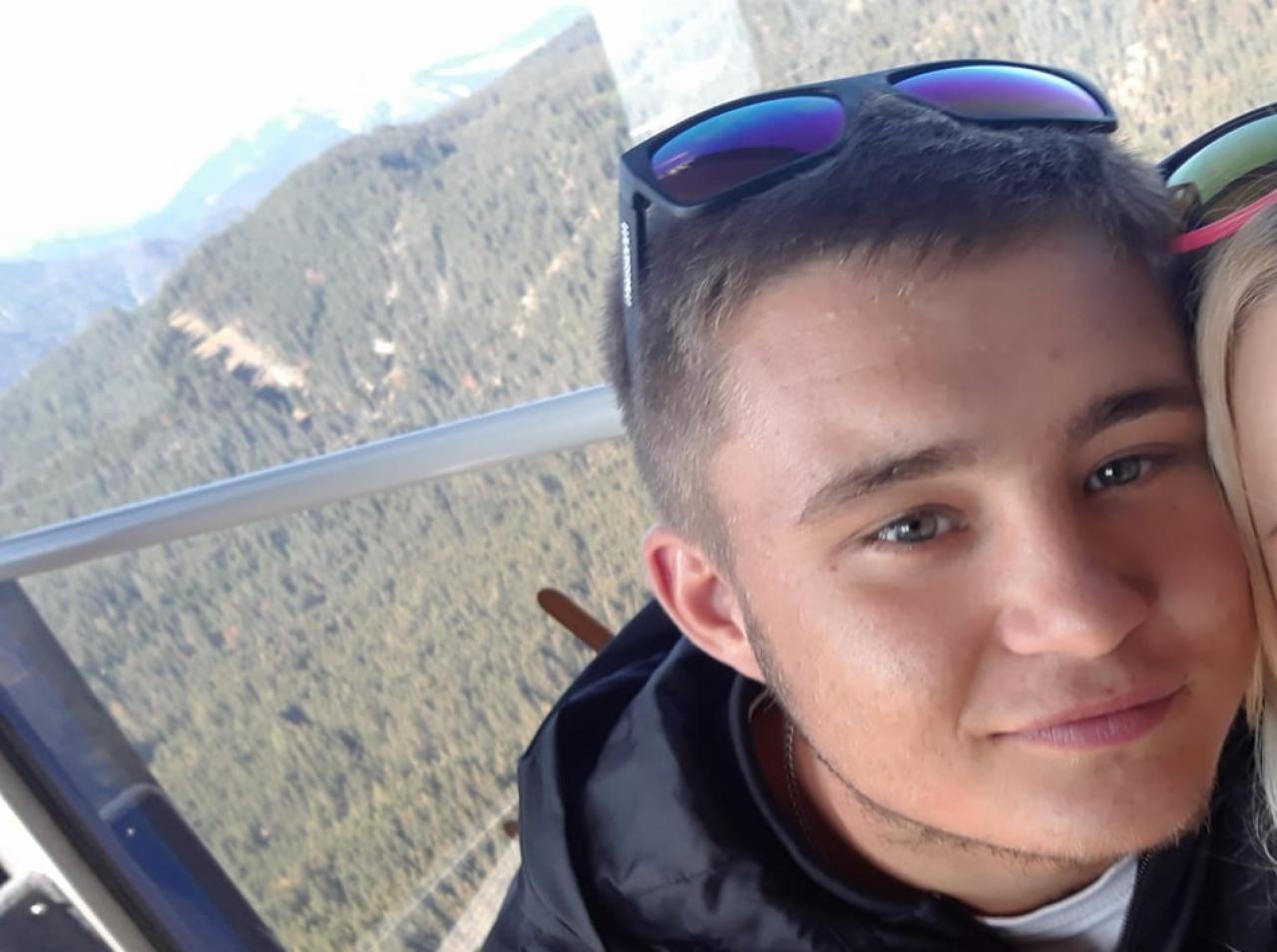 Młody mężczyzna w kurtce i okularach na głowie, robiący selfie na tle zalesionych gór widzianych z okna pojazdu. Widoczne fragmenty konstrukcji pojazdu i drugiej osoby.