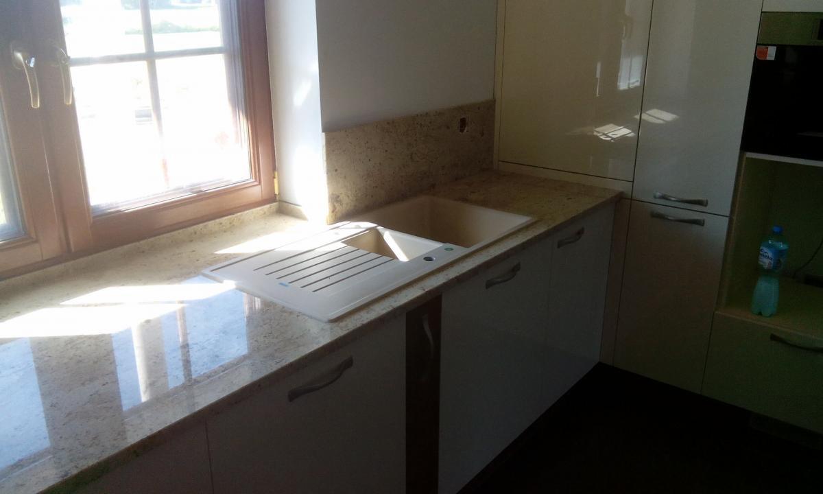 Jasny blat kuchenny z beżowego kamienia z wbudowanym zlewem, widok na okno z drewnianą ramą i część białych szafek kuchennych.