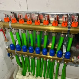 Rozdzielacz ogrzewania podłogowego z zielonymi rurami, zamontowany na ścianie z bloczków, zabezpieczony pianką montażową.