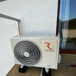 Zewnętrzna jednostka klimatyzacji Rotenso IMOTO, zamontowana na czarnych podkładkach antywibracyjnych, widoczna przy białej ścianie budynku i oknie z odbiciem nieba.