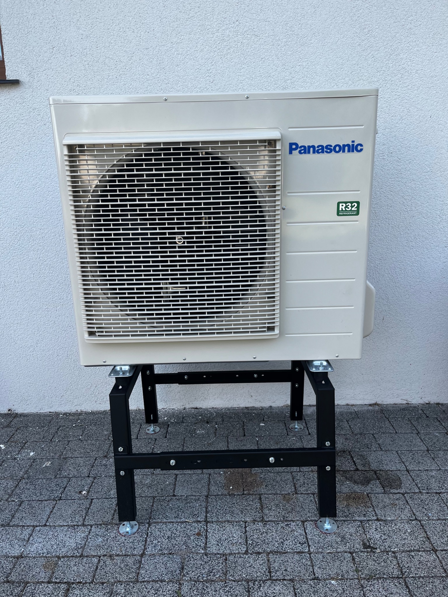 Biała pompa ciepła Panasonic R32 na czarnym metalowym stelażu z regulowanymi nóżkami, ustawiona na szarym bruku przed jasną ścianą.