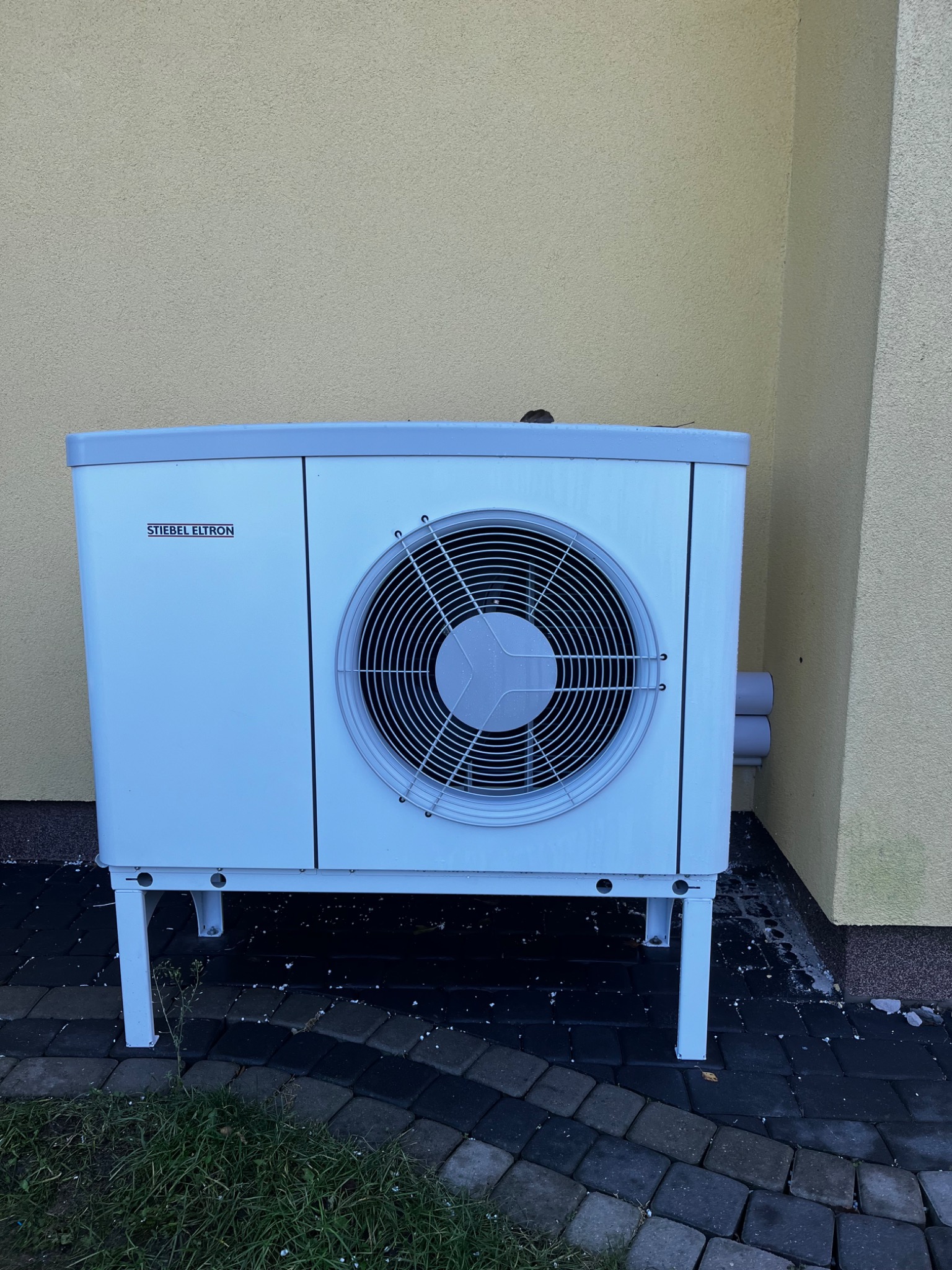 Biała pompa ciepła marki Stiebel Eltron, umieszczona na kostce brukowej przy żółtej ścianie budynku. Widoczne rury przyłączeniowe i fragment trawnika.