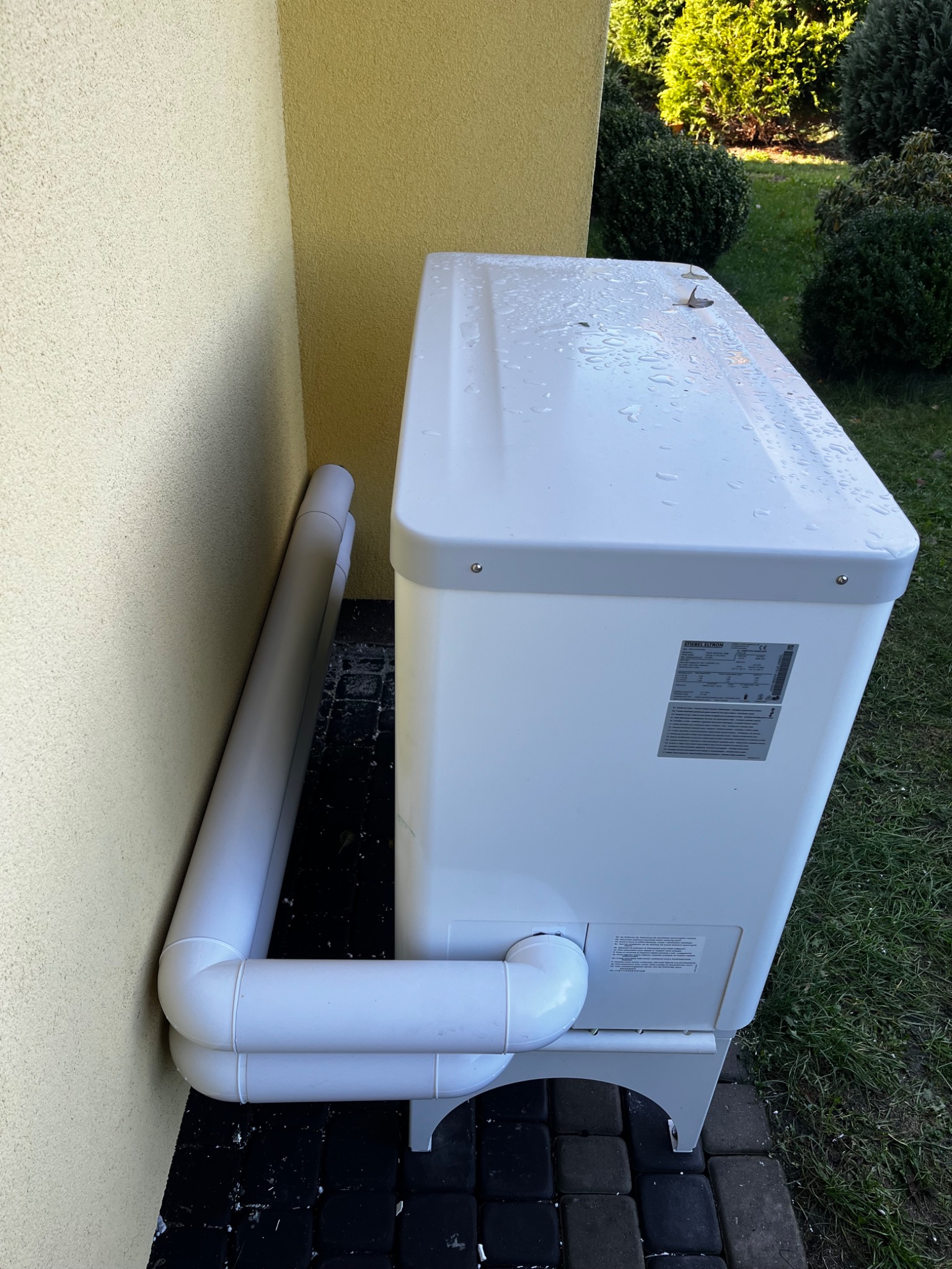 Biała pompa ciepła Stiebel Eltron zamontowana przy elewacji budynku, widoczne rury doprowadzające, krople wody na obudowie i tabliczka znamionowa.