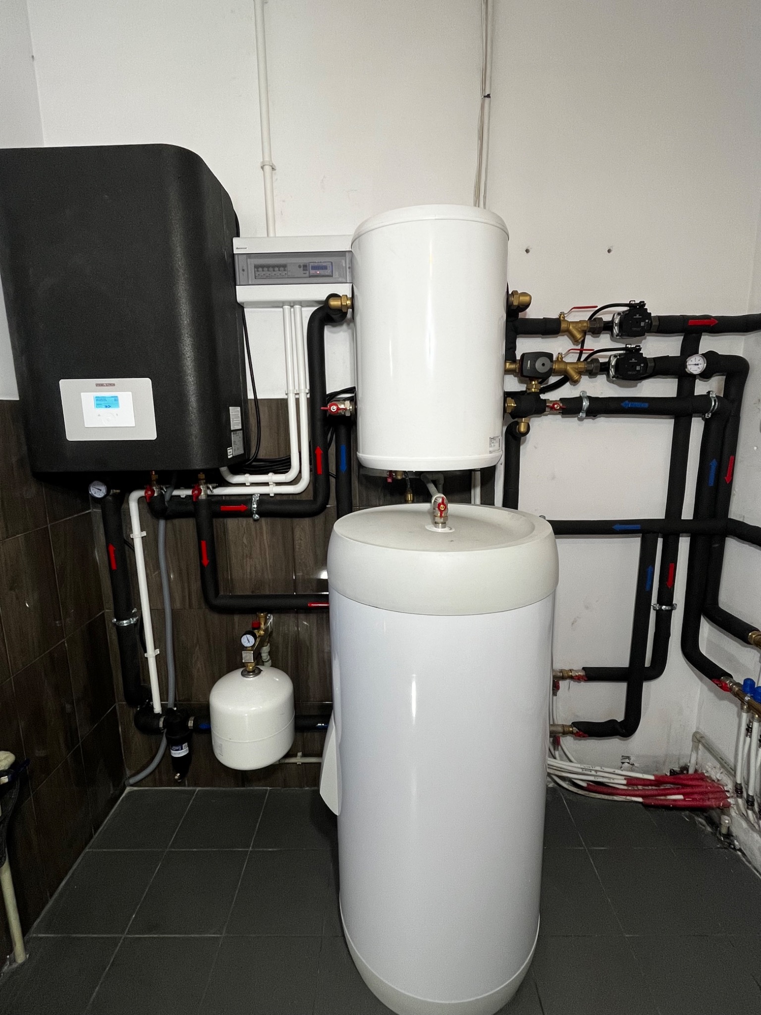 Instalacja grzewcza w kotłowni: czarny zbiornik buforowy obok dwóch białych akumulatorów ciepła połączonych rurami z zaworami i manometrami, widoczny panel sterowania na ścianie.