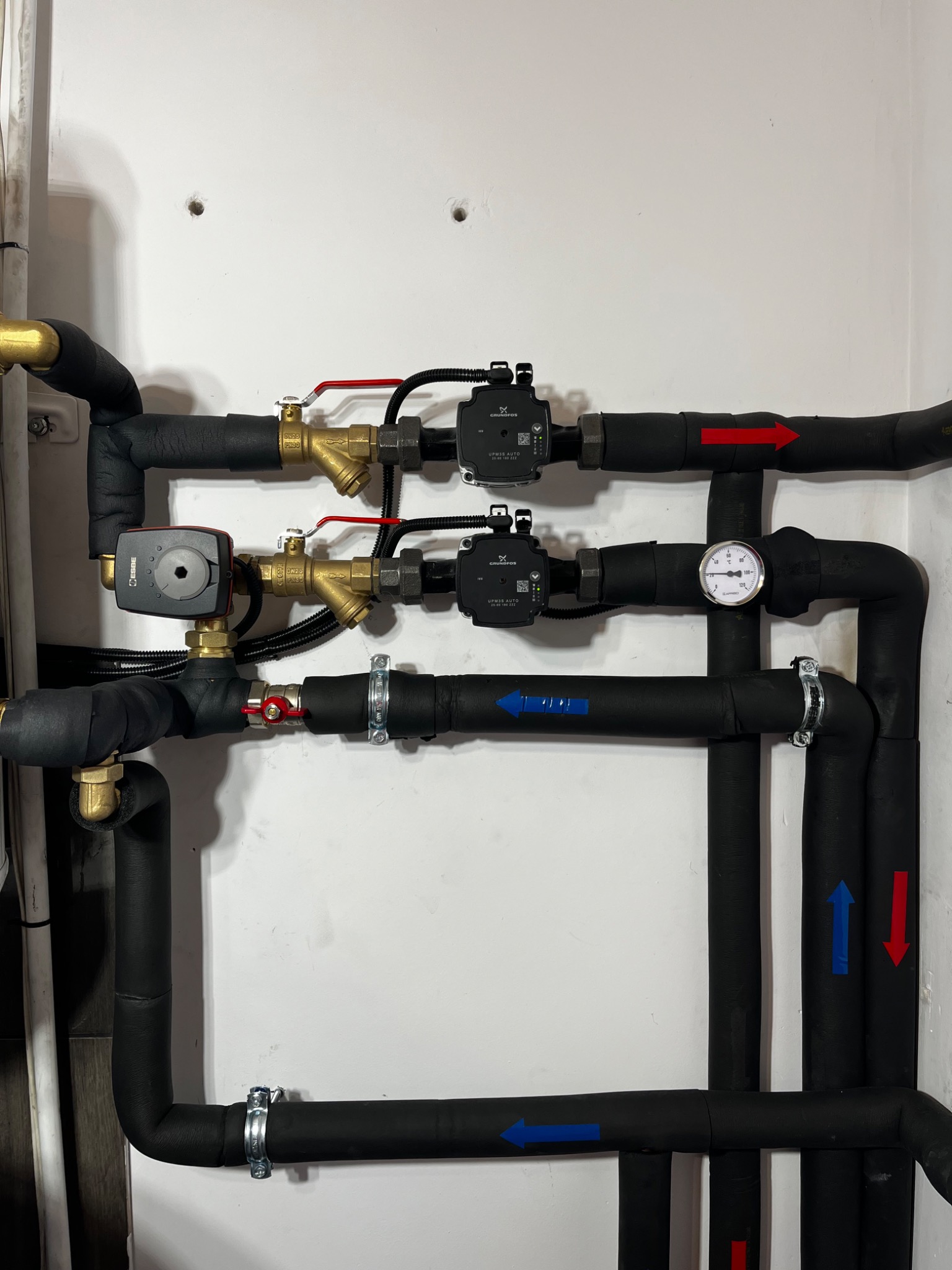 Fragment instalacji grzewczej z dwoma pompami obiegowymi Grundfos, zaworami odcinającymi i manometrem, rury zaizolowane czarną otuliną, strzałki wskazują kierunek przepływu.