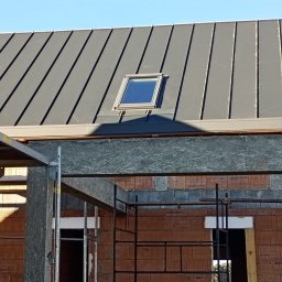 Nivo - Szary dach z blachy na budynku w trakcie budowy, widoczne rusztowania i okno dachowe. Ujęcie z dołu, błękitne niebo w tle.