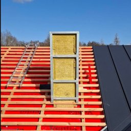 Nivo - Dach w trakcie ocieplania: widoczna konstrukcja z izolacją wełnianą, czerwona membrana, łaty, drabina i fragment pokrycia z blachy na tle błękitnego nieba.