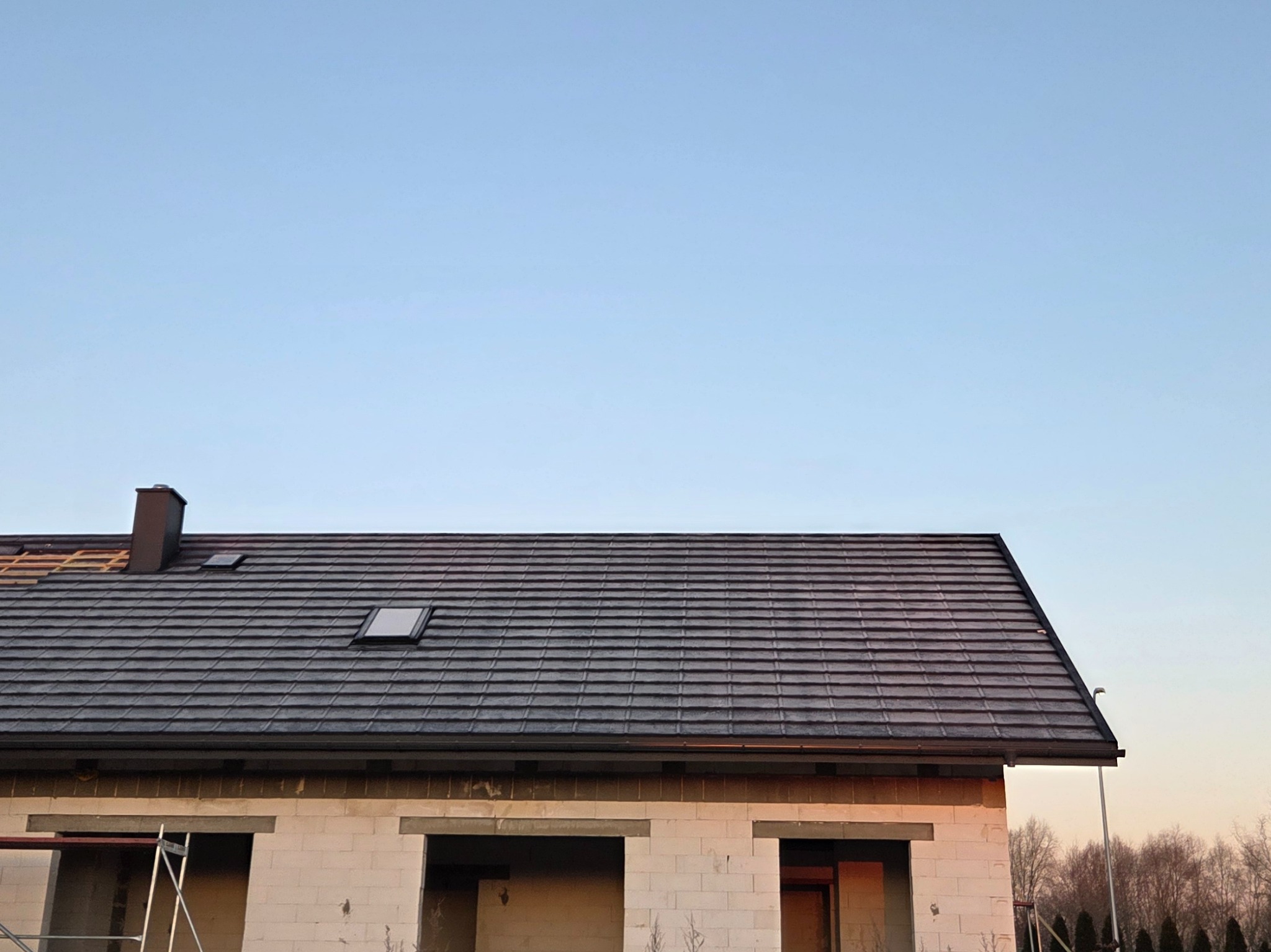 Dom w trakcie budowy z częściowo pokrytym dachem szarą dachówką. Widoczne okno dachowe i komin. Część dachu jeszcze bez pokrycia.