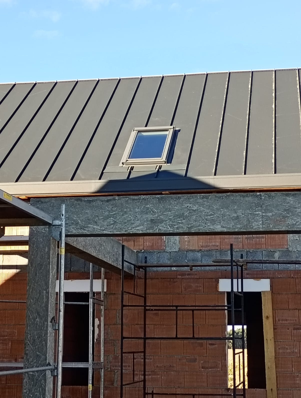 Szary dach z blachy na budynku w trakcie budowy, widoczne rusztowania i okno dachowe. Ujęcie z dołu, błękitne niebo w tle.