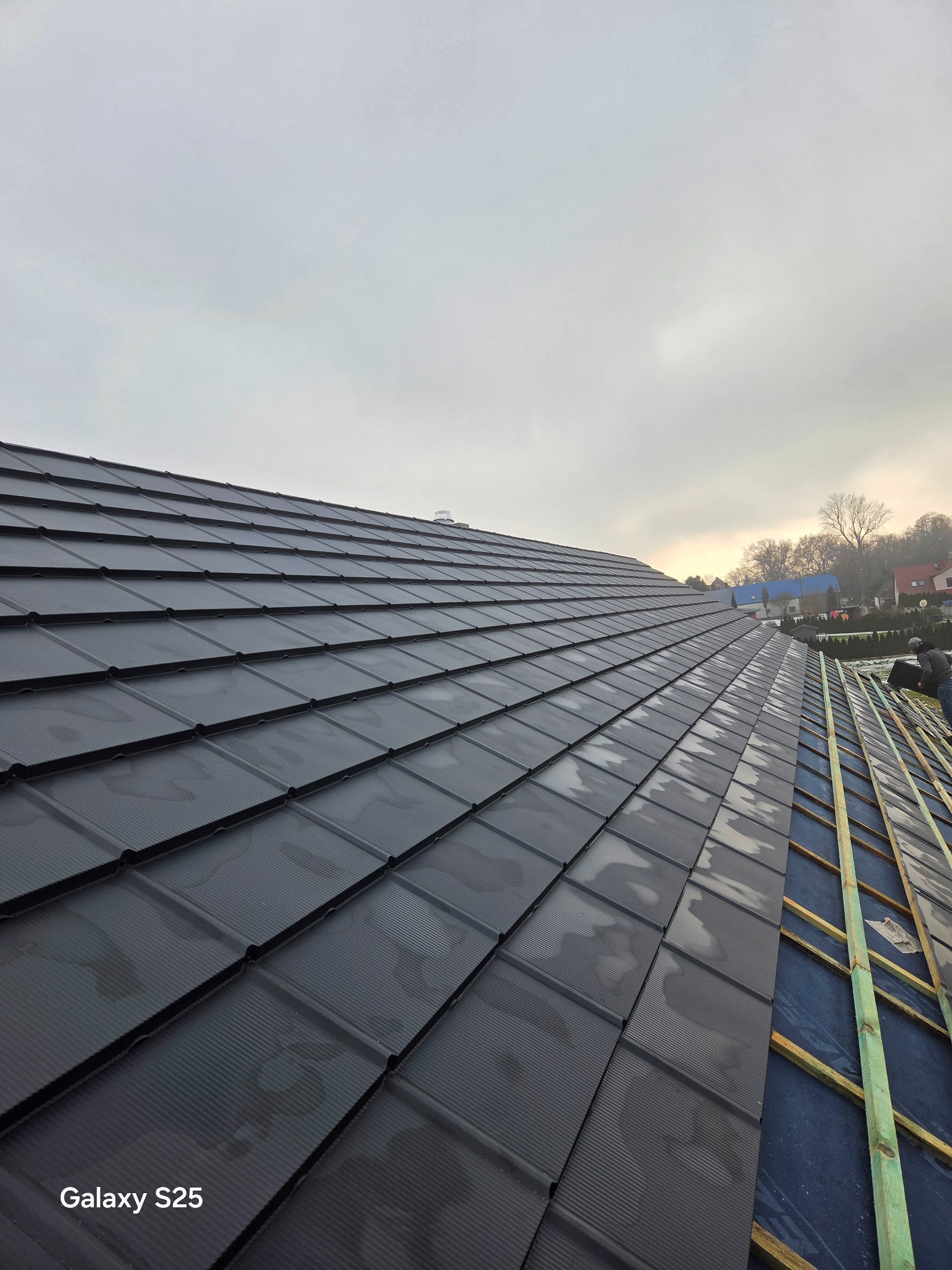 Częściowo pokryty dach z czarnej blachodachówki modułowej, widoczne łaty i membrana. W tle zabudowa jednorodzinna i pochmurne niebo. Ujęcie z dołu.