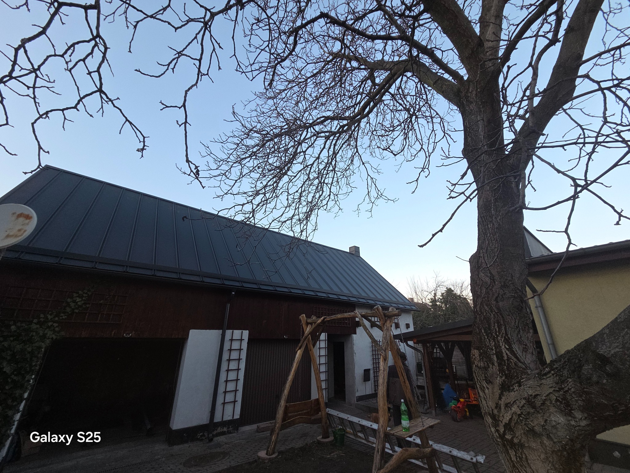 Dom z ciemnym, metalowym dachem widziany z dołu, z fragmentem drzewa z przodu. Widoczne rynny i elementy konstrukcyjne dachu. Ujęcie z niskiej perspektywy.