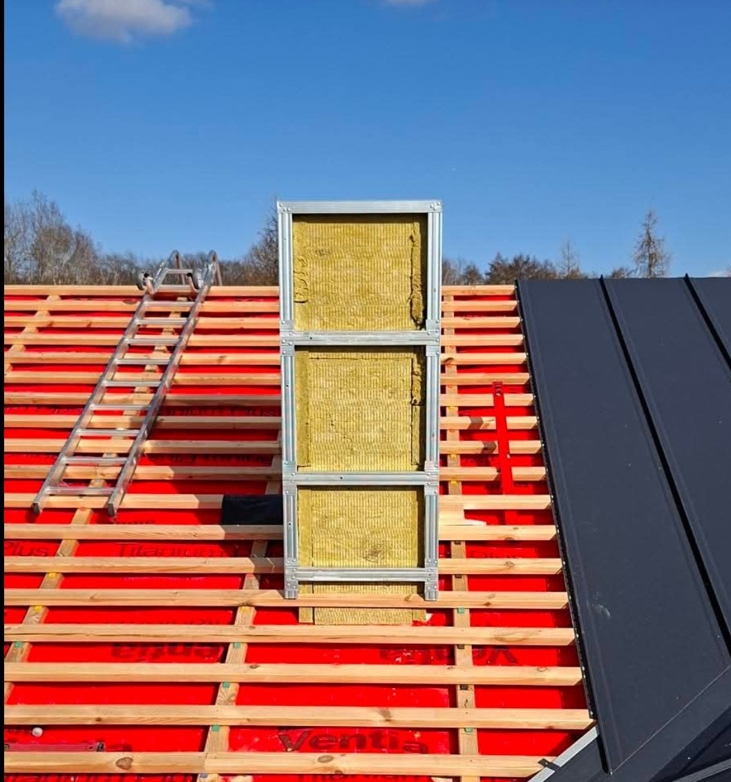 Dach w trakcie ocieplania: widoczna konstrukcja z izolacją wełnianą, czerwona membrana, łaty, drabina i fragment pokrycia z blachy na tle błękitnego nieba.