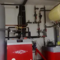 Kompleksowa kotłownia z dwoma czerwonymi piecami marki Brastal, zasobnikiem wody i rozbudowaną instalacją rur miedzianych i czarnych, widok na urządzenia od frontu w jasnym pomieszczeniu.