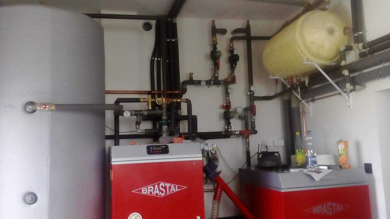 Kompleksowa kotłownia z dwoma czerwonymi piecami marki Brastal, zasobnikiem wody i rozbudowaną instalacją rur miedzianych i czarnych, widok na urządzenia od frontu w jasnym pomieszczeniu.