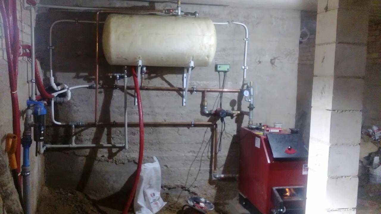 Instalacja kotła CO z zasobnikiem w kotłowni, widoczne rury miedziane i plastikowe, filtr wody, ściany z betonu komórkowego, na podłodze rozsypana ziemia.
