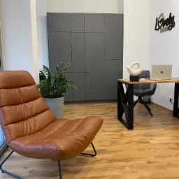 Jasne biuro z brązowym skórzanym fotelem, szarą szafą w zabudowie, drewnianym biurkiem i czarnym krzesłem biurowym. Na ścianie dekoracja 'Lovely house'.