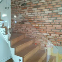 Drewniane schody z balustradą ze szkła i białymi elementami mocującymi na tle ceglanej ściany, widoczne przewody elektryczne i miarka.