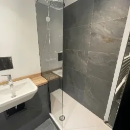 Nowoczesna łazienka z kabiną prysznicową typu walk-in, umywalką nablatową na drewnianym blacie i szarymi płytkami imitującymi kamień.