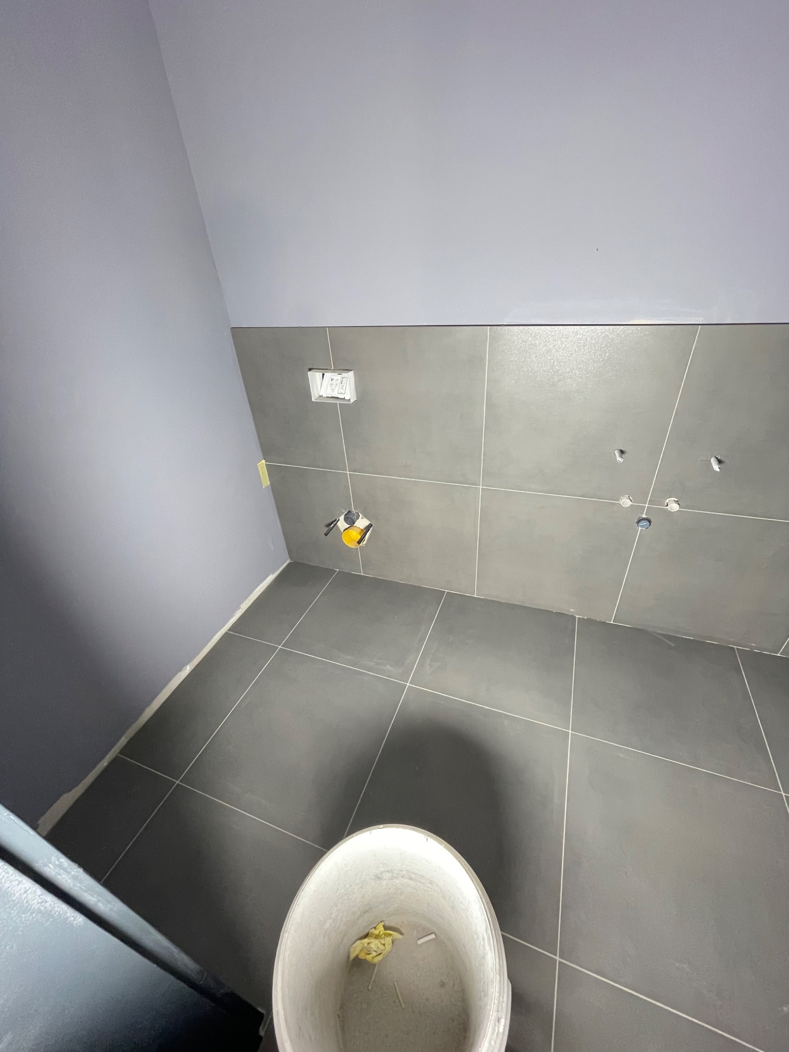 Szare płytki podłogowe i ścienne w trakcie układania, widoczne otwory na instalacje, wiadro z resztkami zaprawy w rogu pomieszczenia.