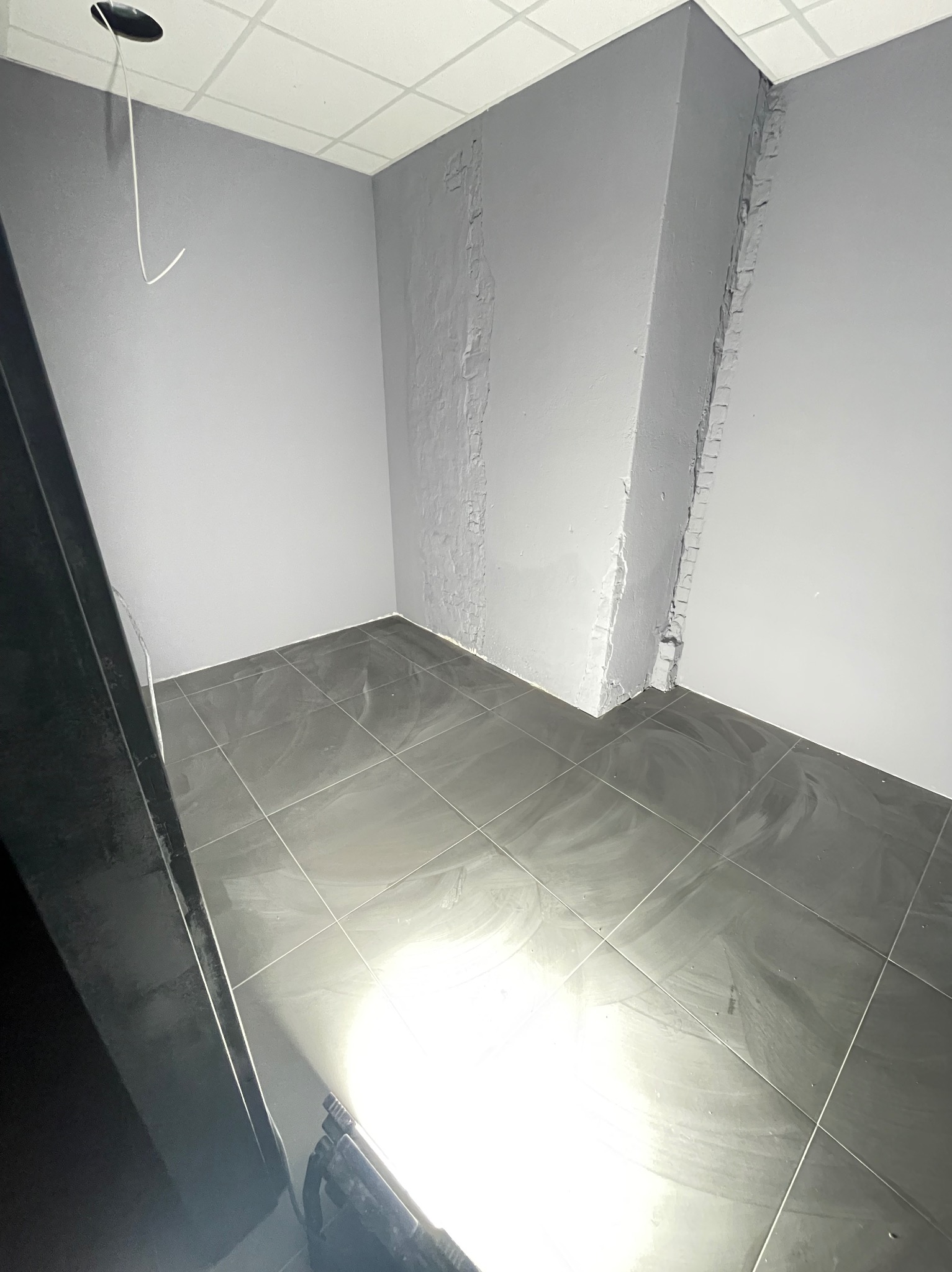 Szare płytki podłogowe z widocznymi śladami po czyszczeniu w rogu pomieszczenia z niedokończonymi ścianami i wystającym przewodem z sufitu.