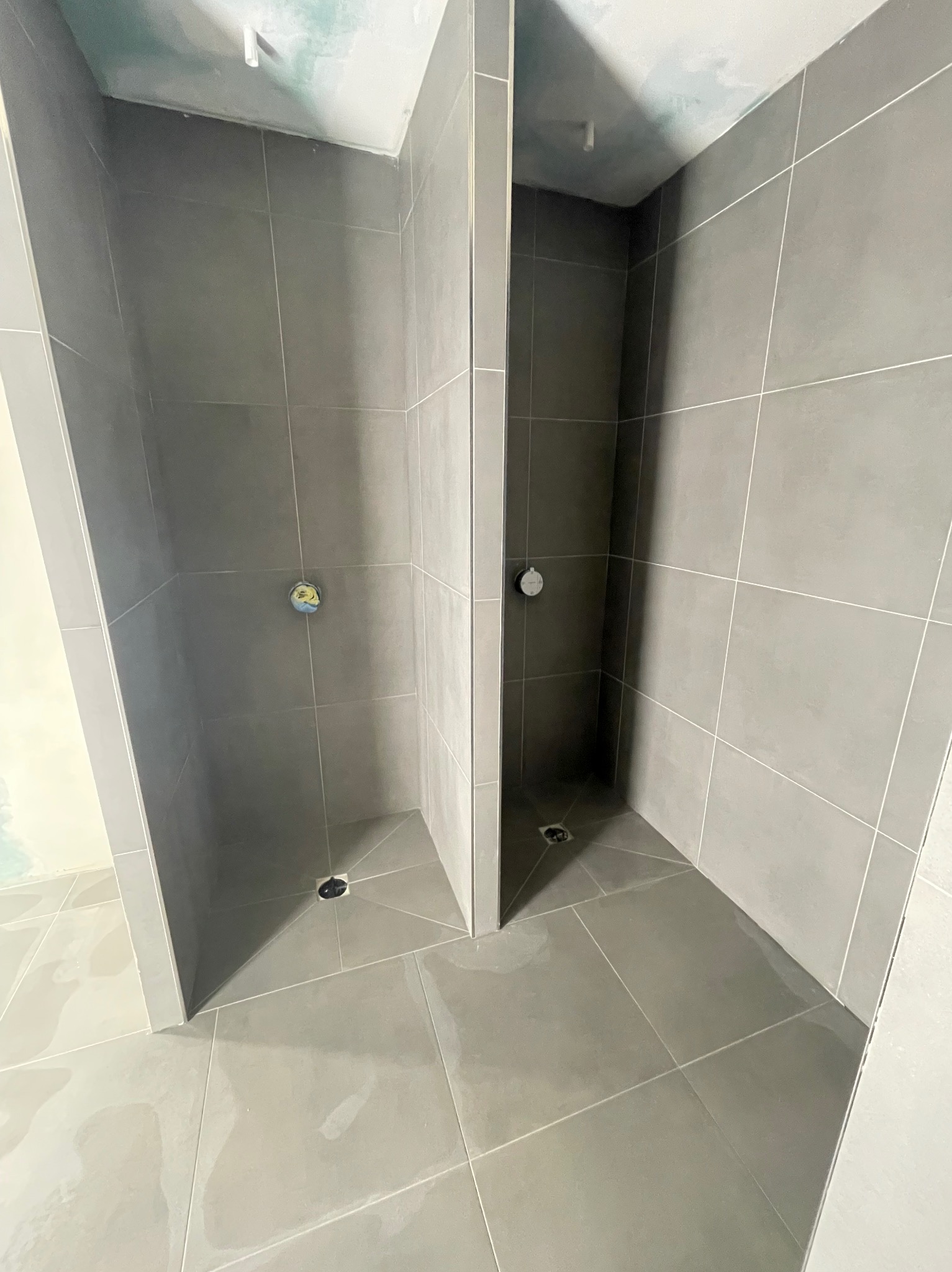 Dwie kabiny prysznicowe w trakcie wykańczania, wyłożone szarymi płytkami, widoczne odpływy liniowe i wystające rury instalacji wodnej.