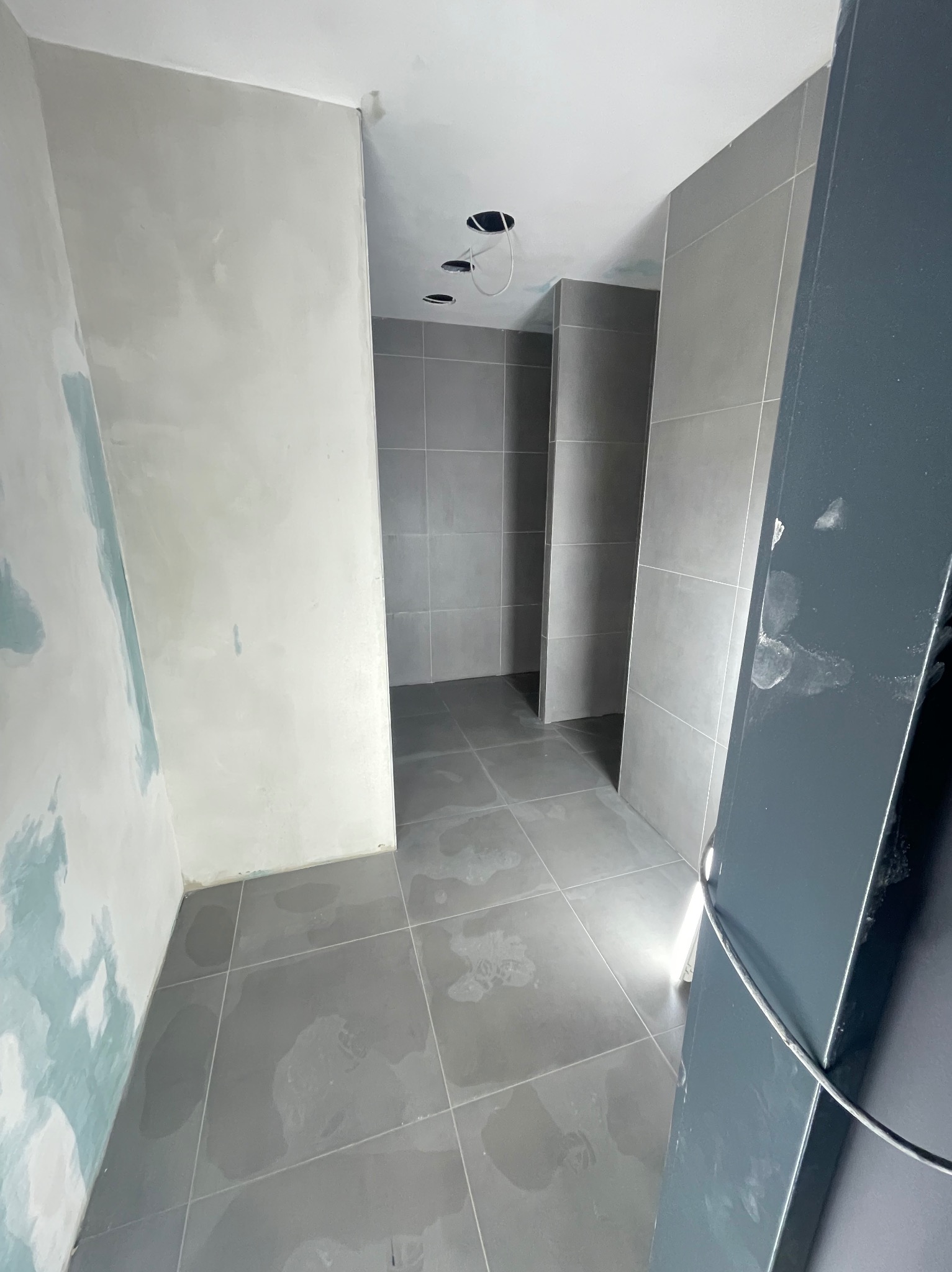 Wnętrze łazienki w trakcie remontu: szare płytki na podłodze i ścianach, ściana w trakcie szpachlowania, widoczne otwory na oświetlenie sufitowe.