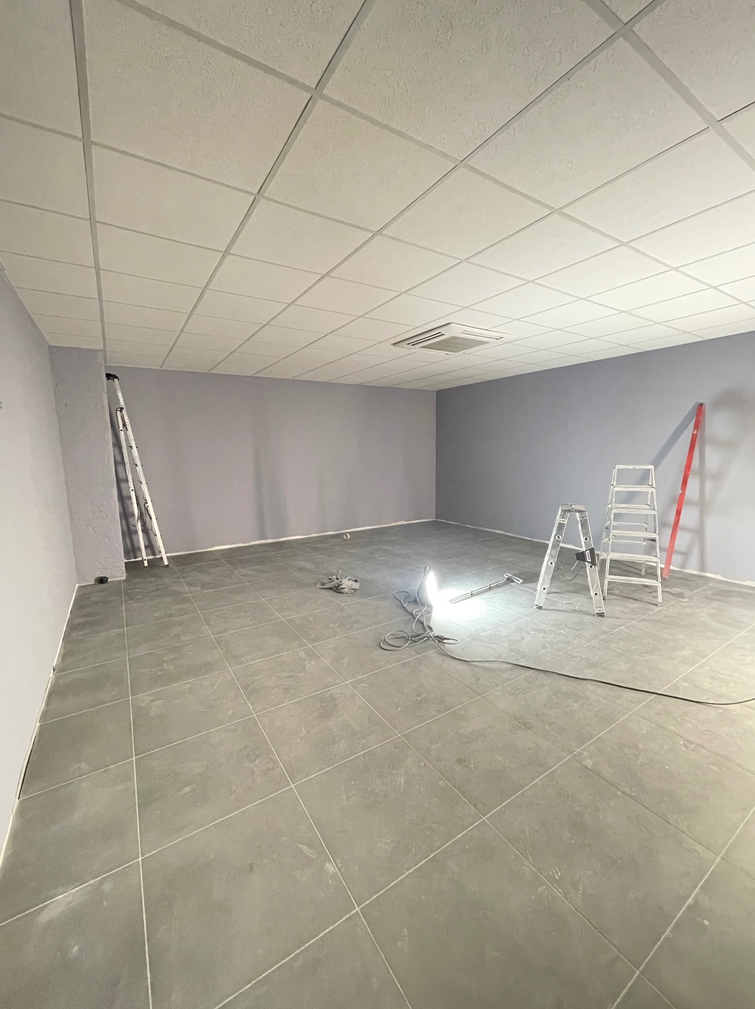 Pomieszczenie w trakcie malowania ścian na szaro, z widocznymi drabinami, lampą roboczą i podwieszanym sufitem kasetonowym.