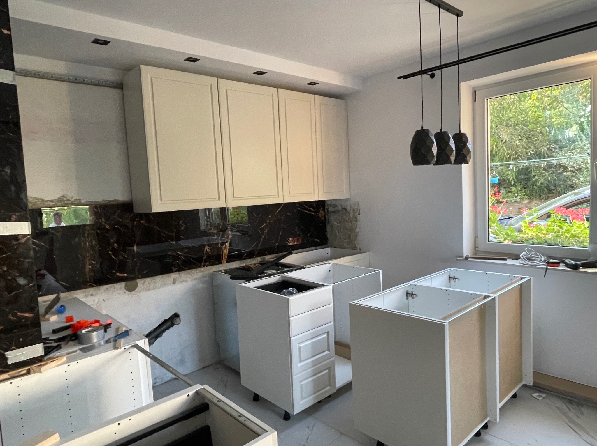 Montaż szafek kuchennych z kremowymi frontami i czarnym marmurowym panelem nad blatem, widoczne narzędzia i elementy montażowe.