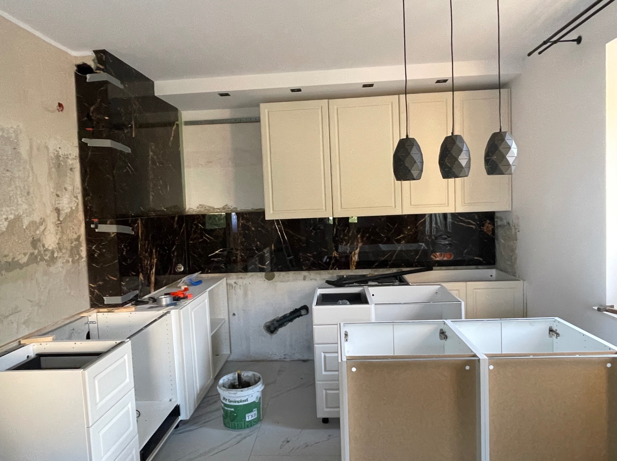 Montaż kuchni: widoczne niedokończone szafki dolne z białymi frontami i górne z kremowymi, czarny panel nad blatem imitujący marmur, trzy wiszące lampy o geometrycznych kloszach, ściany...