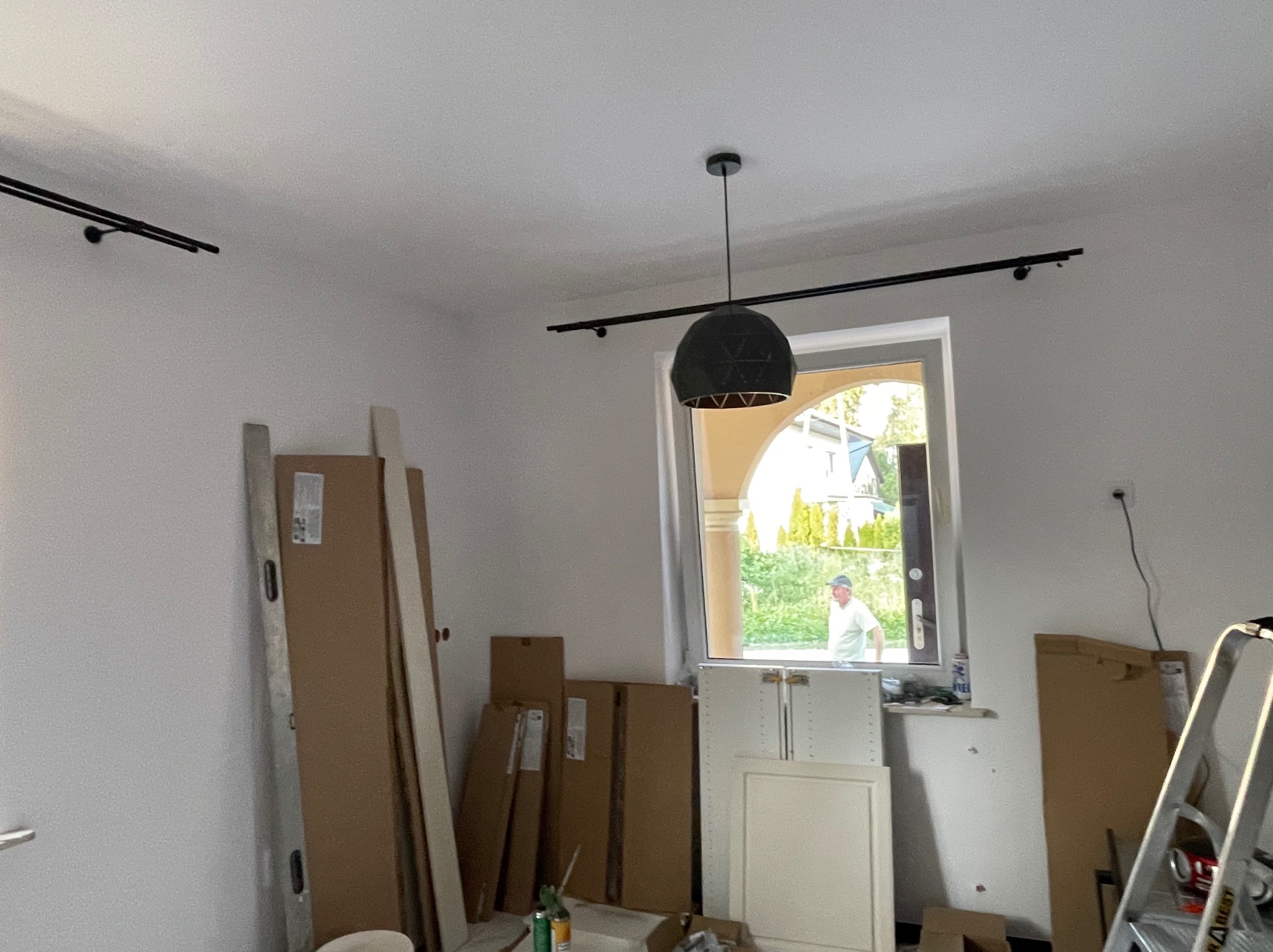 Wnętrze w trakcie wykańczania: czarna lampa wisząca, kartony, drabina, poziomica, widok na zewnątrz przez okno z kolumnami.