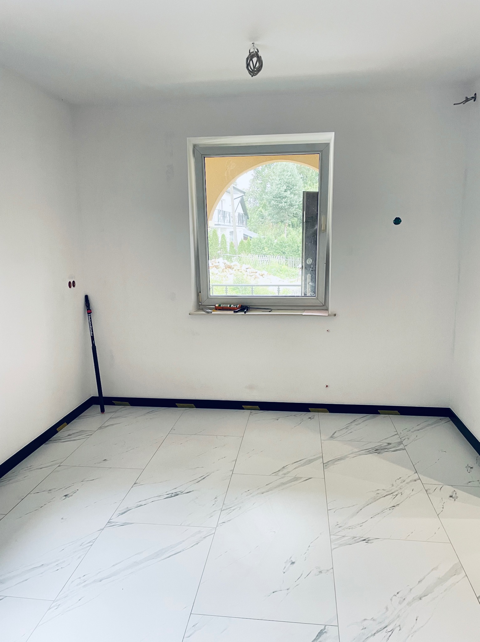 Pusty pokój z białymi ścianami, marmurową podłogą i oknem z widokiem na zieleń, widoczne narzędzia na parapecie i listwy przypodłogowe jeszcze zabezpieczone taśmą.
