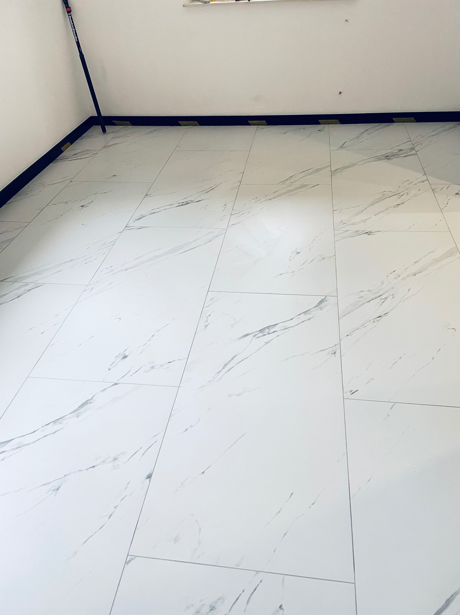 Białe, marmurowe płytki podłogowe ułożone w prostokątnym wzorze, zabezpieczone taśmą malarską przy czarnej listwie przypodłogowej, widoczne narzędzie malarskie w rogu.
