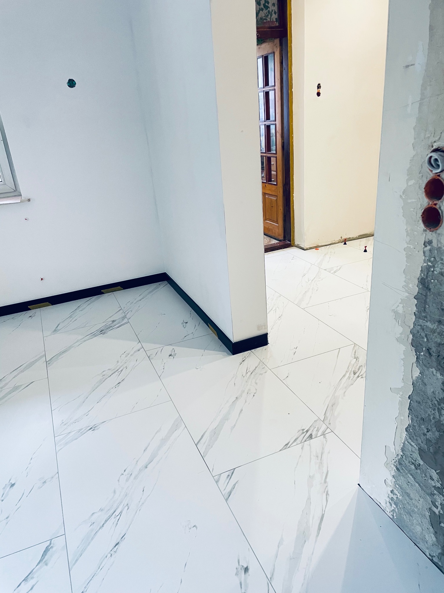 Ułożona podłoga z białych płytek imitujących marmur, widoczne czarne listwy przypodłogowe, niedokończone ściany i otwór drzwiowy z drewnianymi drzwiami w tle.
