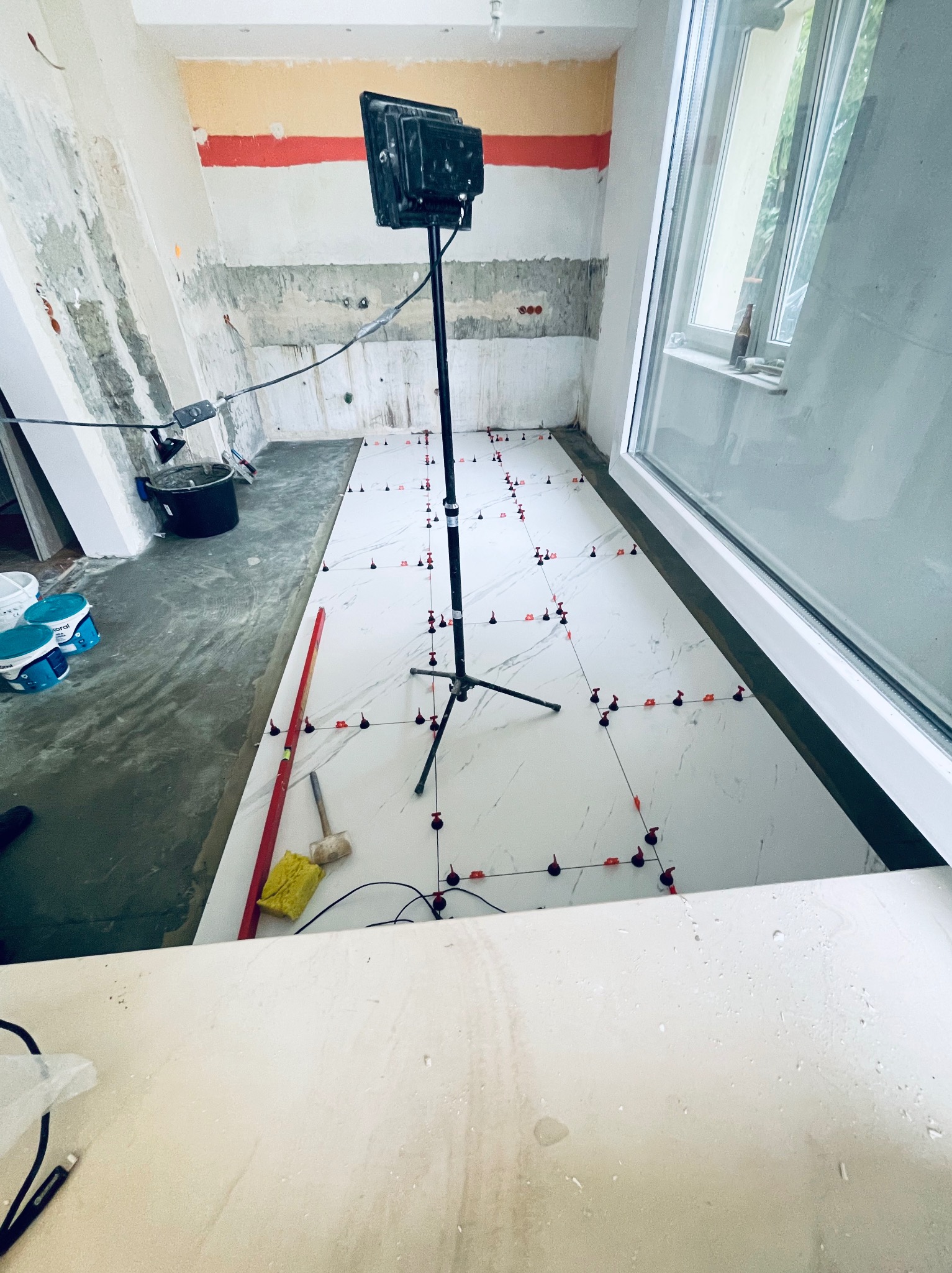 Układanie białych płytek imitujących marmur w pomieszczeniu w trakcie remontu, widoczne poziomice, młotek gumowy, krzyżyki dystansowe i lampa budowlana.