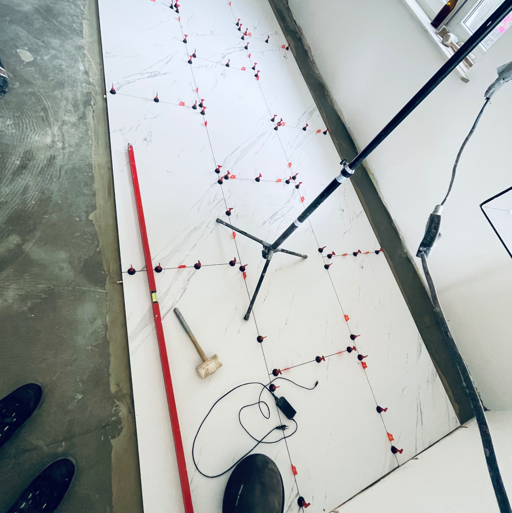 Układanie białych płytek imitujących marmur na podłodze z użyciem poziomicy, młotka gumowego i systemu poziomowania płytek z czerwonymi klipsami, widoczne buty glazurnika i statyw oświetleniowy