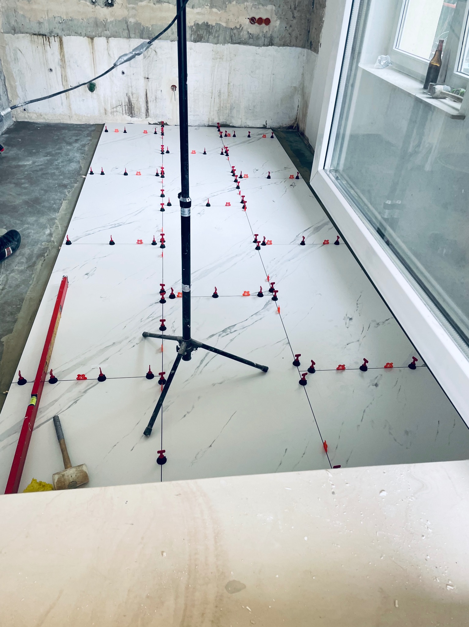 Układanie białych płytek imitujących marmur na podłodze, z użyciem systemu poziomowania płytek (klipsy i kliny), widoczny poziomica, młotek gumowy i fragment ściany w stanie surowym.