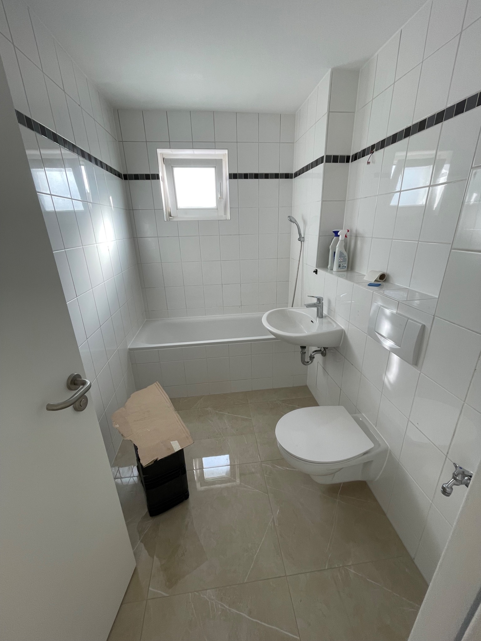 Wnętrze łazienki w trakcie remontu, z widoczną wanną, umywalką i toaletą, wykończone białymi płytkami z czarnym akcentem, na podłodze beżowe płytki imitujące marmur, widoczne pudełko z kartonem...