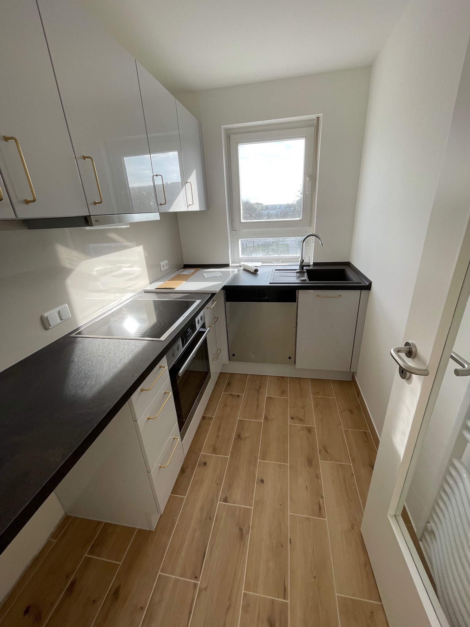 Nowoczesna, mała kuchnia w odcieniach bieli i beżu z czarnym blatem, złotymi uchwytami i drewnopodobną podłogą z płytek, oświetlona światłem dziennym wpadającym przez okno.