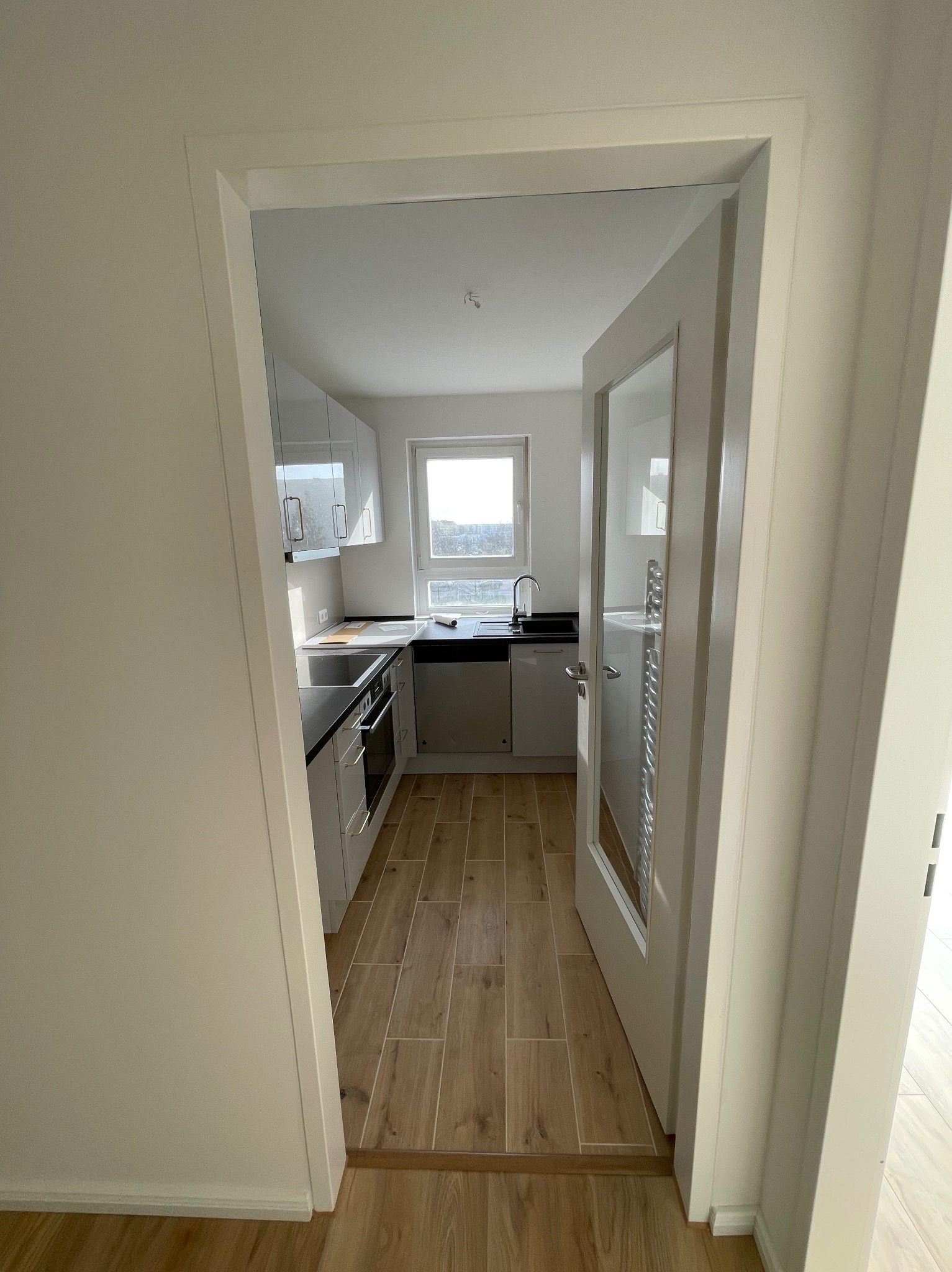 Widok przez otwarte drzwi na nową, jasną kuchnię z drewnopodobną podłogą, białymi szafkami i czarnym blatem.