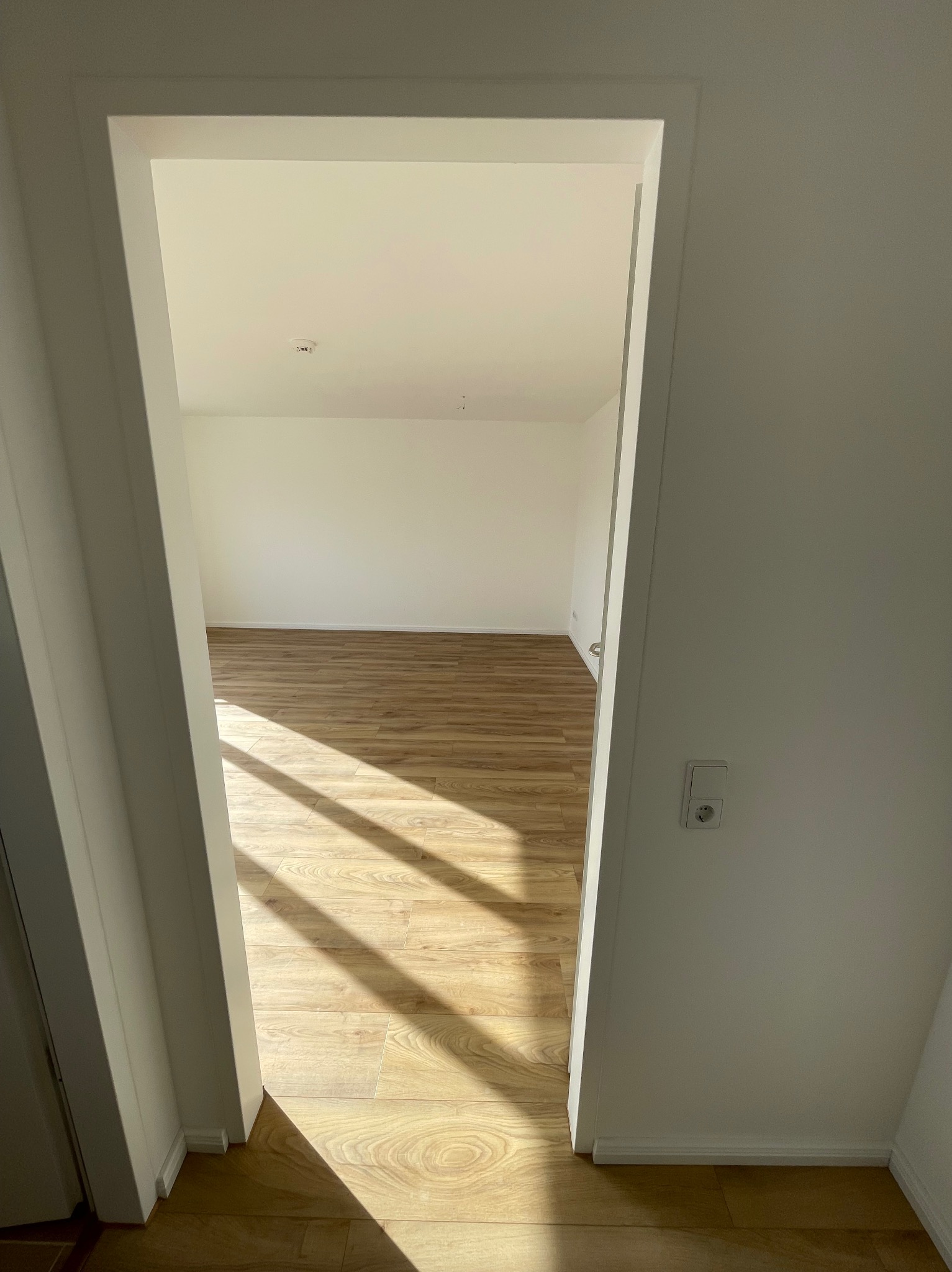 Widok przez białe futryny drzwi na puste, jasne pomieszczenie z drewnianą podłogą, oświetlone promieniami słońca, z widocznym gniazdkiem elektrycznym na ścianie.