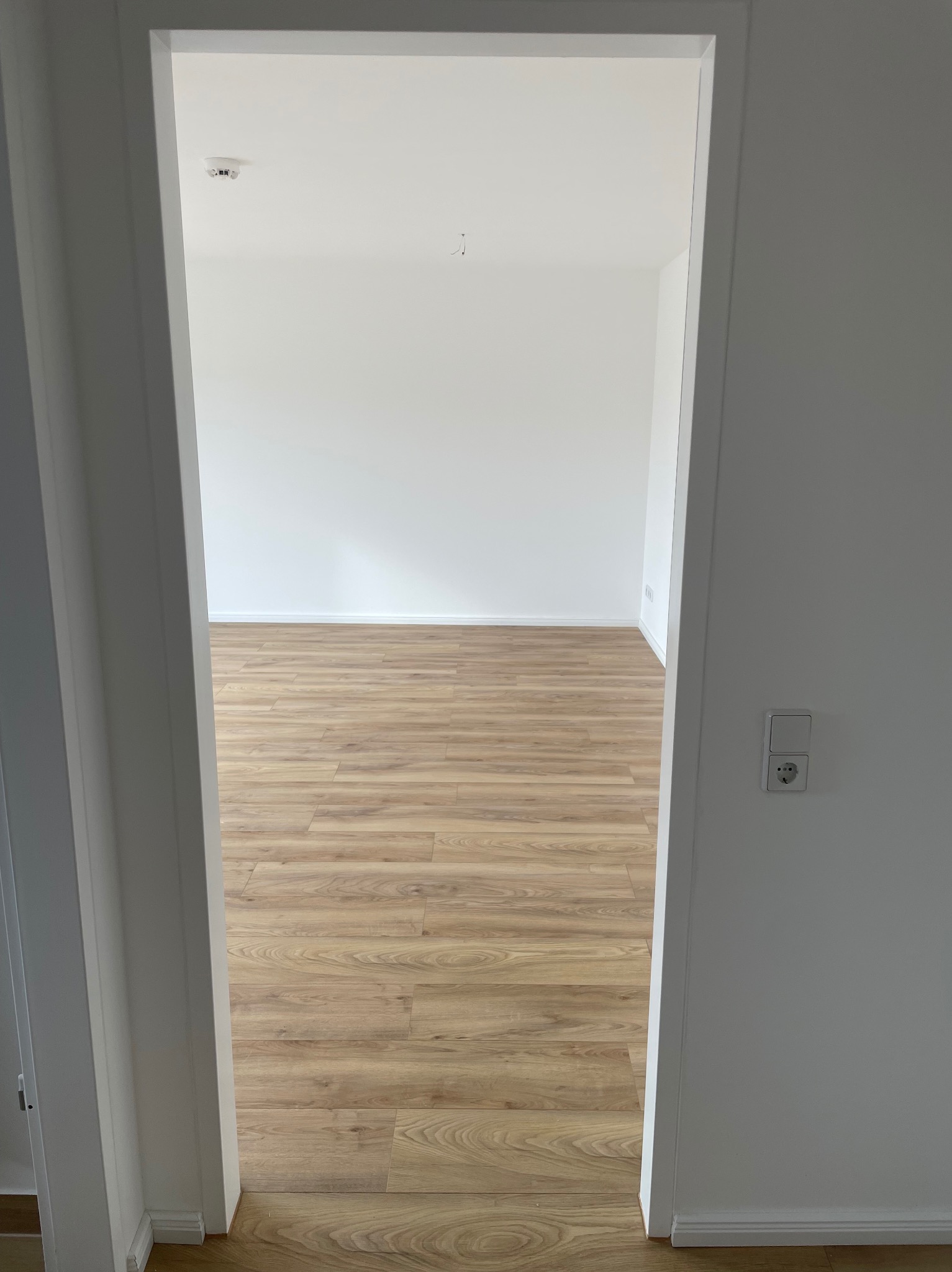 Widok przez białe drzwi na puste, wykończone wnętrze z panelami podłogowymi w odcieniach brązu i beżu, białe ściany z widocznym gniazdkiem elektrycznym i czujnikiem dymu na suficie.