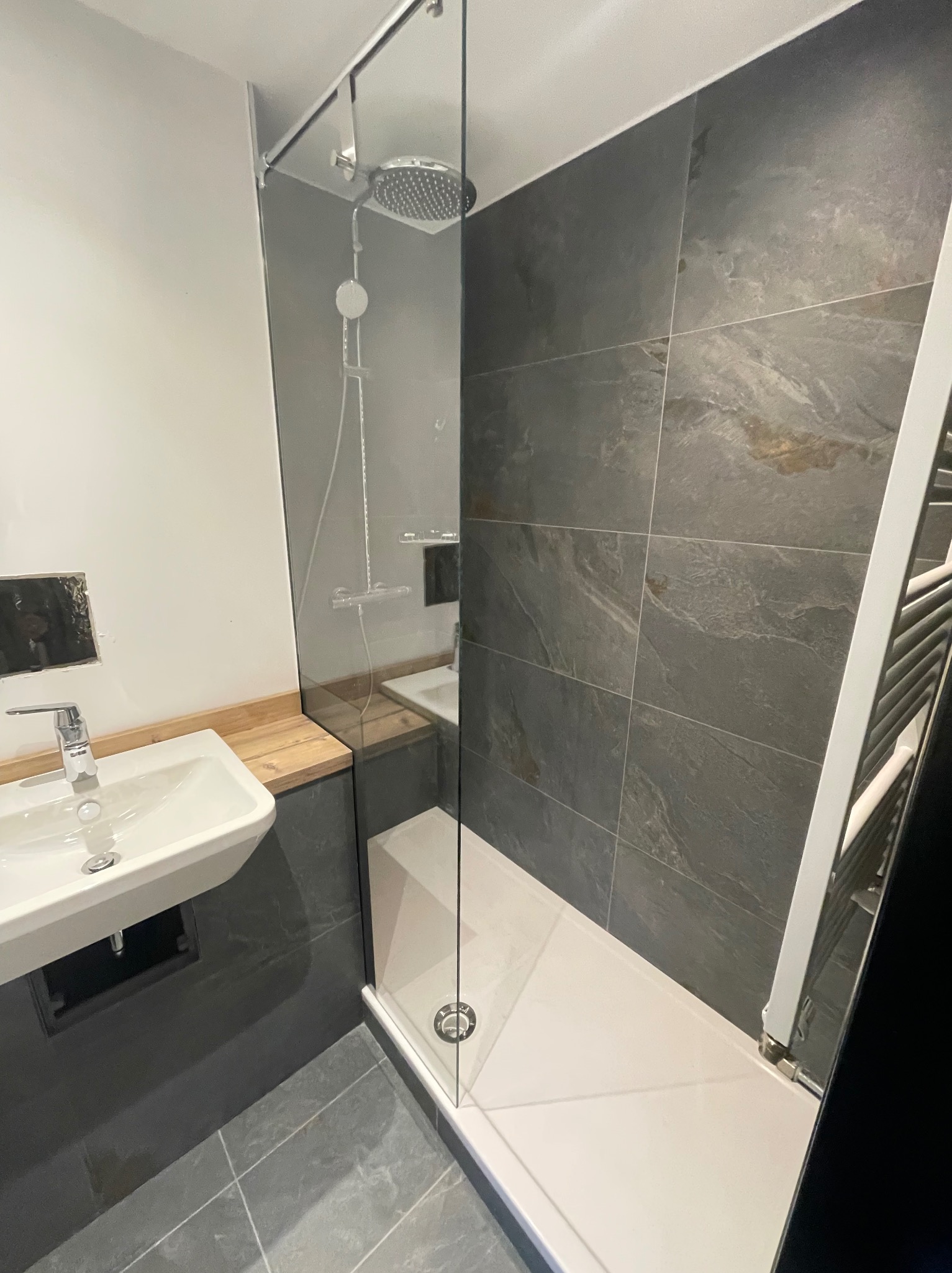 Nowoczesna łazienka z kabiną prysznicową typu walk-in, umywalką nablatową na drewnianym blacie i szarymi płytkami imitującymi kamień.