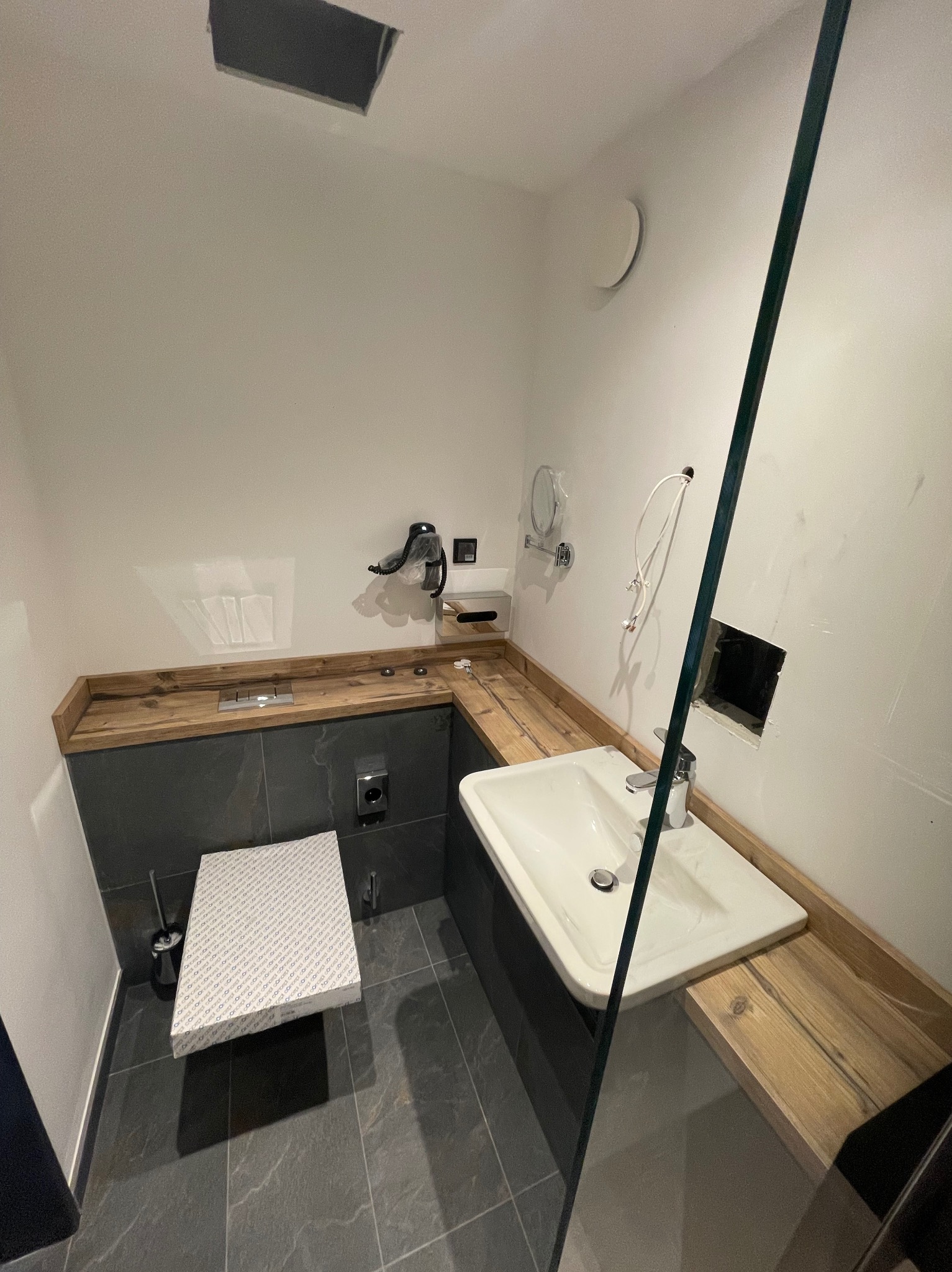 Łazienka w trakcie remontu: biała umywalka z drewnianym blatem, toaleta w opakowaniu, widoczne niedokończone instalacje i otwory w ścianie.