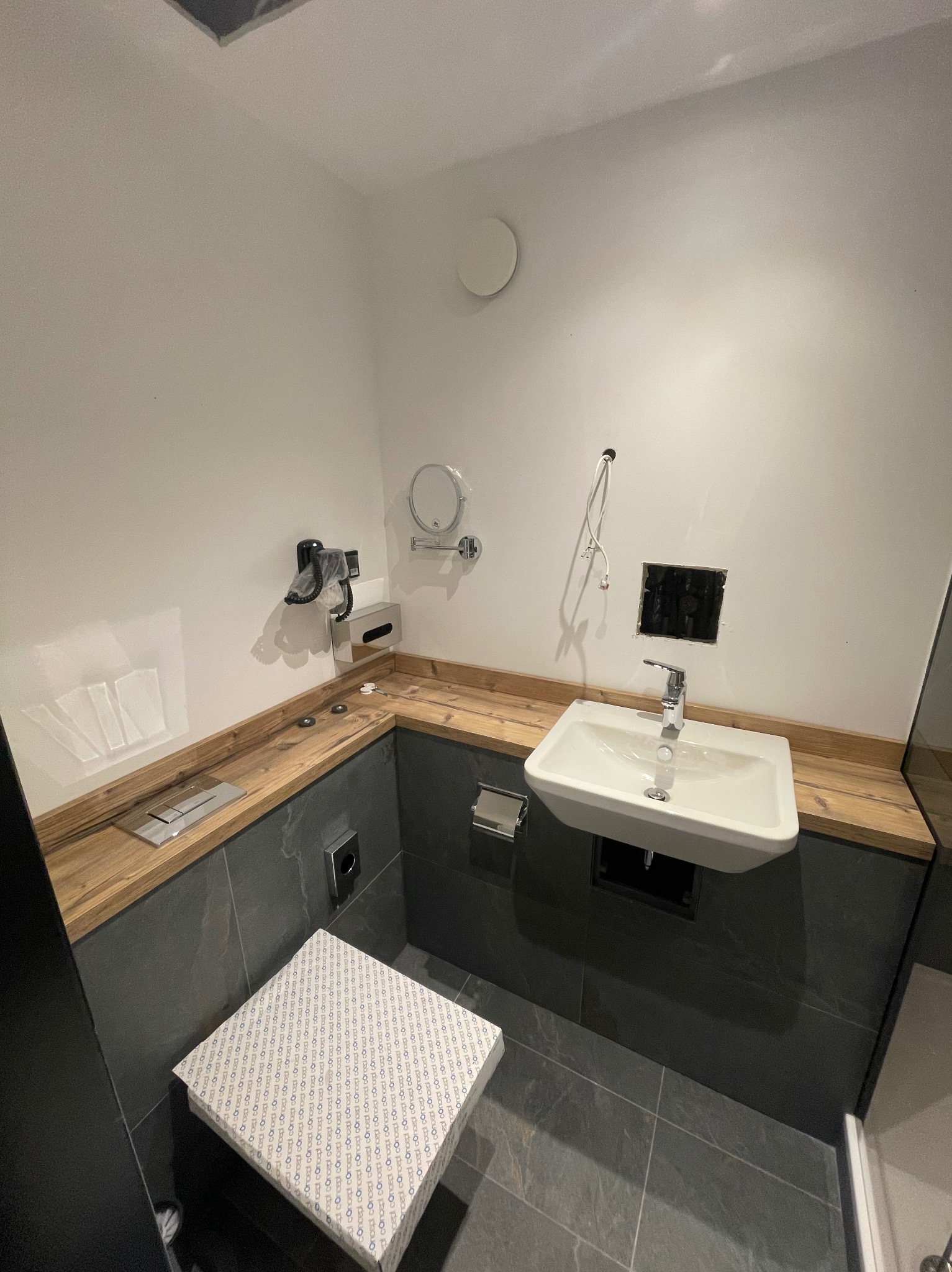 Łazienka w trakcie remontu: biała umywalka zamontowana na drewnianym blacie, widoczne niedokończone podłączenia, obok zapakowana w karton toaleta z nadrukowanym wzorem.