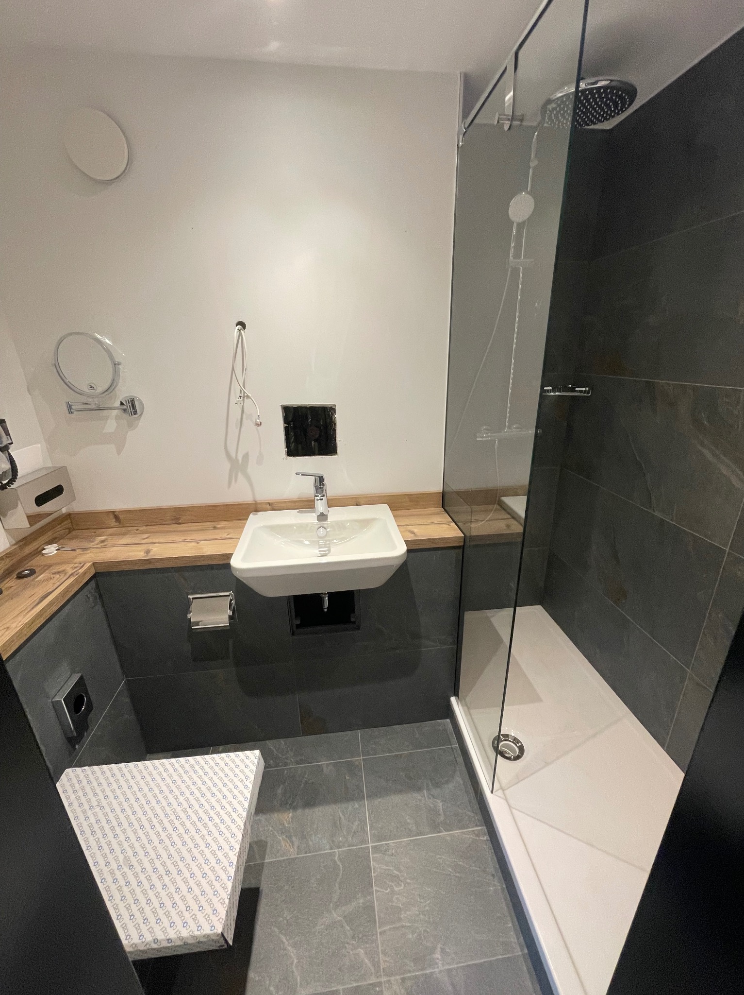 Nowoczesna łazienka w trakcie remontu: biała umywalka nablatowa, szara zabudowa meblowa z drewnianym blatem, szklana kabina prysznicowa z deszczownicą, szare płytki podłogowe, widoczne...