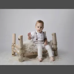 Mały chłopiec w białym ubraniu i szelkach siedzi na łóżeczku z brzozowych bali wyłożonym białym futrem na jasnym tle, machając ręką.