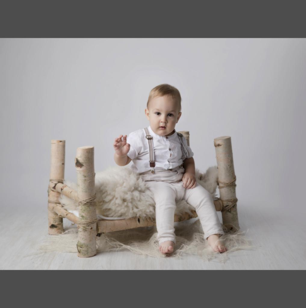 Mały chłopiec w białym ubraniu i szelkach siedzi na łóżeczku z brzozowych bali wyłożonym białym futrem na jasnym tle, machając ręką.