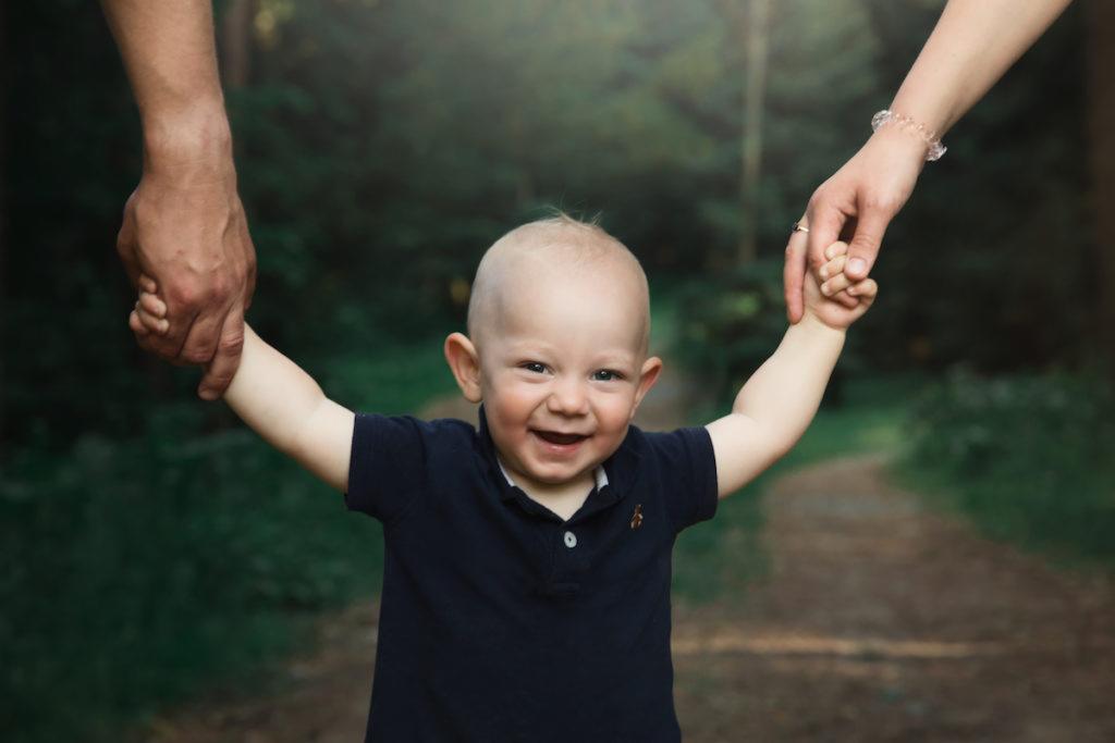 Uśmiechnięty roczny chłopiec w granatowej koszulce polo, trzymany za ręce przez rodziców na leśnej ścieżce, z rozmazanym tłem z drzew.