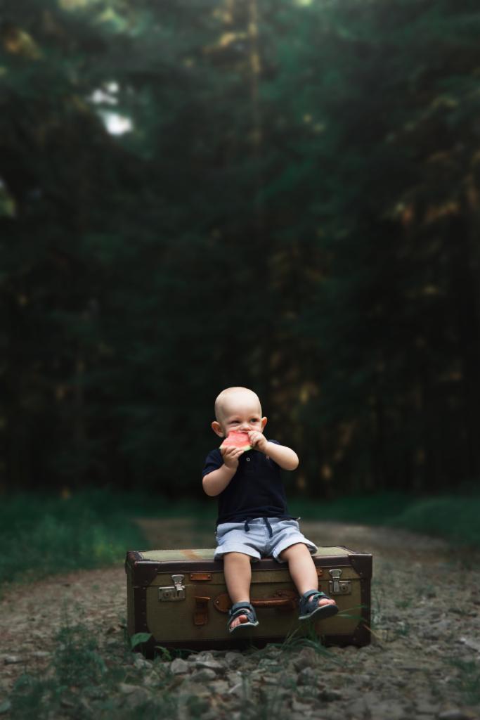 Mały chłopiec siedzi na starej walizce w lesie i je arbuza.