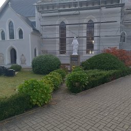 IS-International service - Kościół w trakcie remontu dachu, widoczne rusztowania, część dachu już pokryta nowym materiałem, na pierwszym planie rzeźba na tle zieleni.